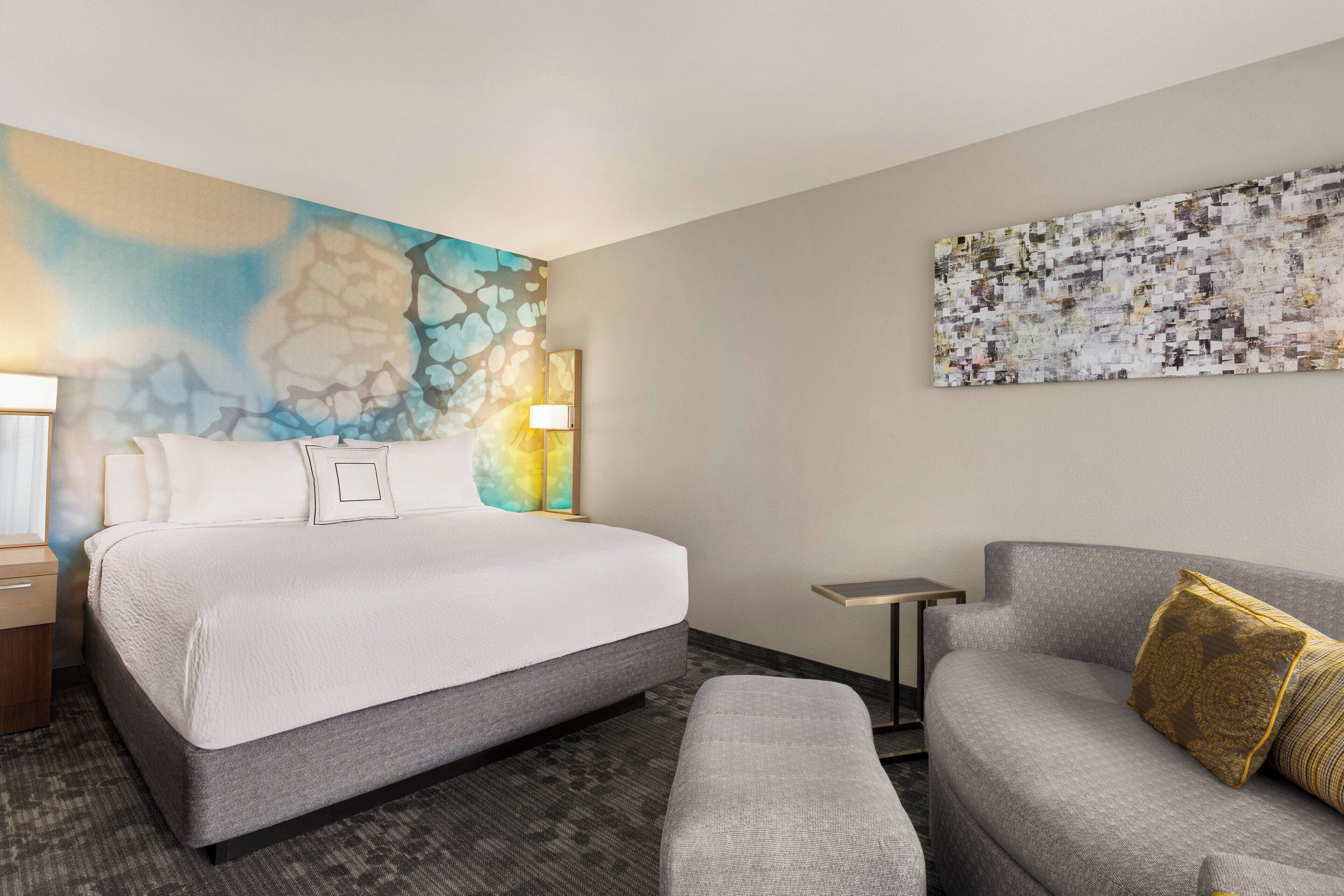 A hotel room with a large bed, a gray sofa, a small table, and two wall art pieces.