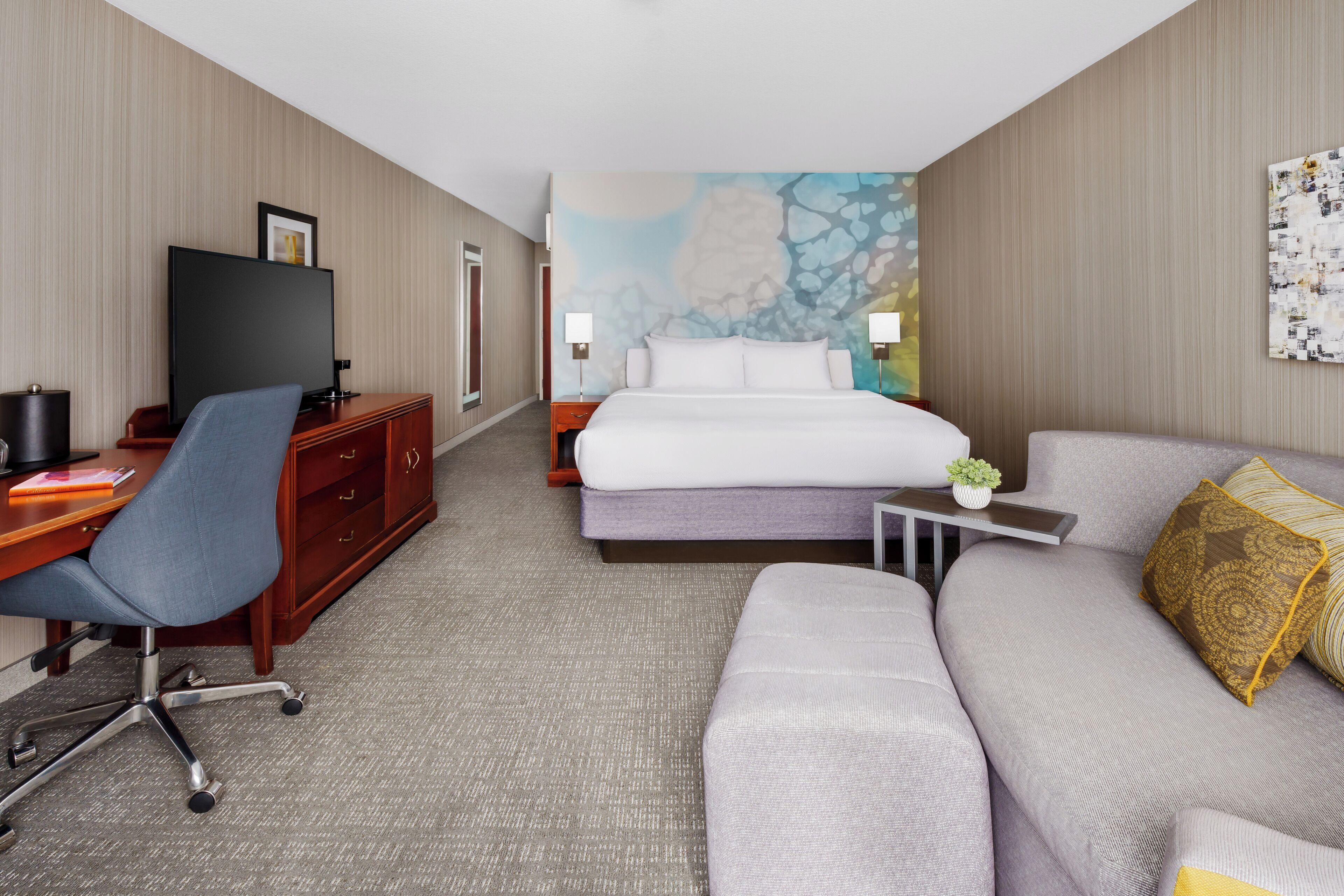 A hotel room with a bed, a desk with a chair, a sofa, a TV, and a wall with a painting.