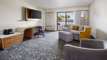 A hotel room with a desk, chair, sofa, ottoman, and a TV mounted on the wall.
