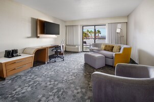 A hotel room with a desk, chair, sofa, ottoman, and a TV mounted on the wall.