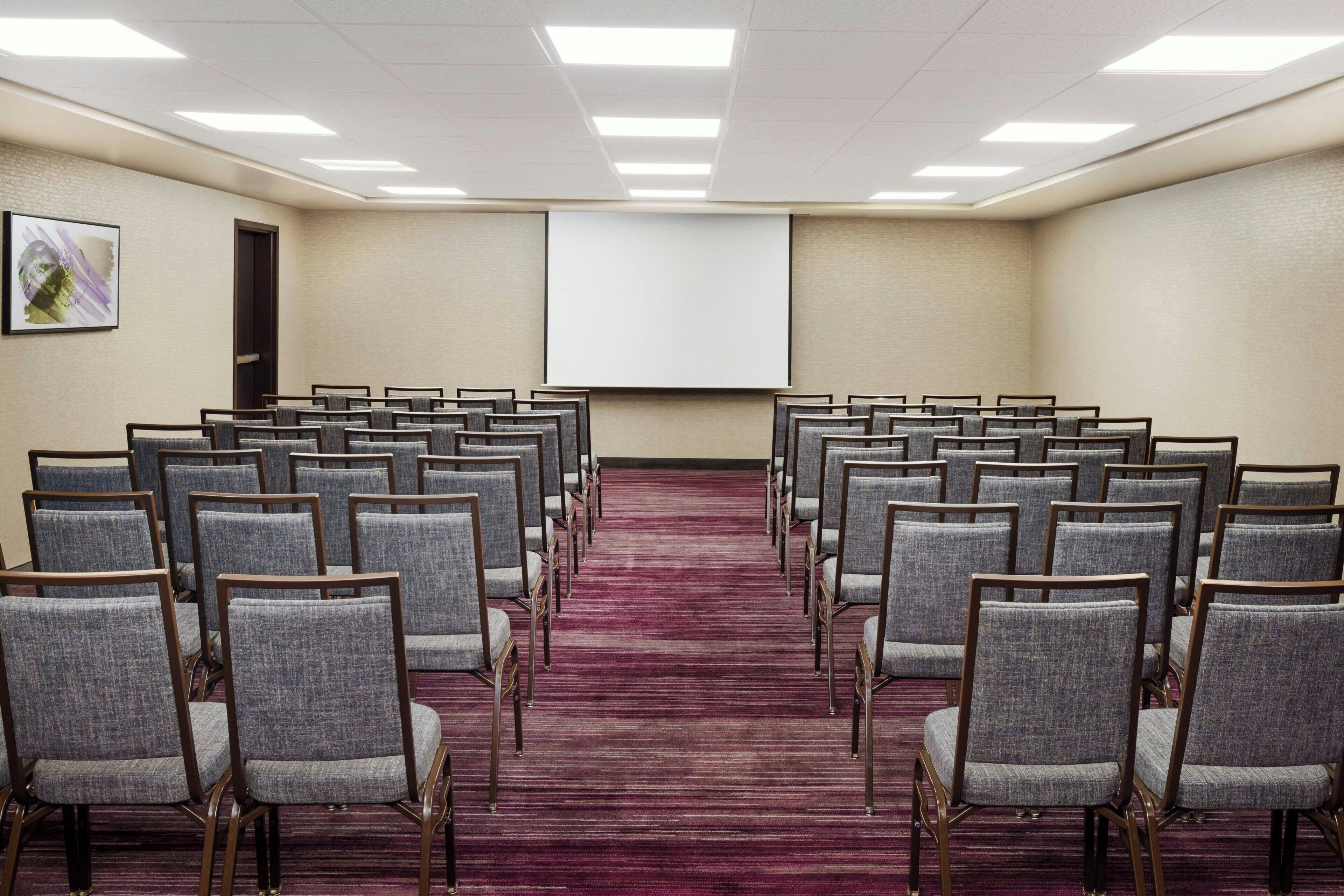 Una sala de conferencias con filas de sillas grises, piso alfombrado, una pantalla de proyector y una puerta.