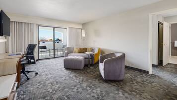 A hotel room with a desk, two armchairs, a sofa, a television, and a large window with a view.