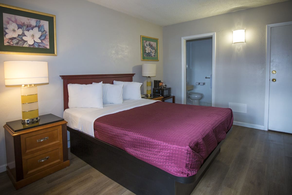 A hotel room with a bed, nightstands, a lamp, a framed floral picture, and a door leading to a bathroom.