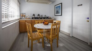 Una pequeña cocina con armarios de madera, una encimera blanca y una mesa redonda con sillas. Hay un cuenco de fruta sobre la mesa y una cafetera en la encimera.