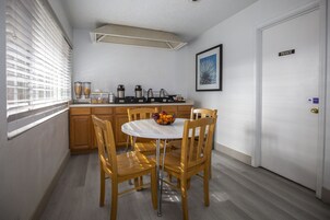 Una pequeña cocina con armarios de madera, una encimera blanca y una mesa redonda con sillas. Hay un cuenco de fruta sobre la mesa y una cafetera en la encimera.