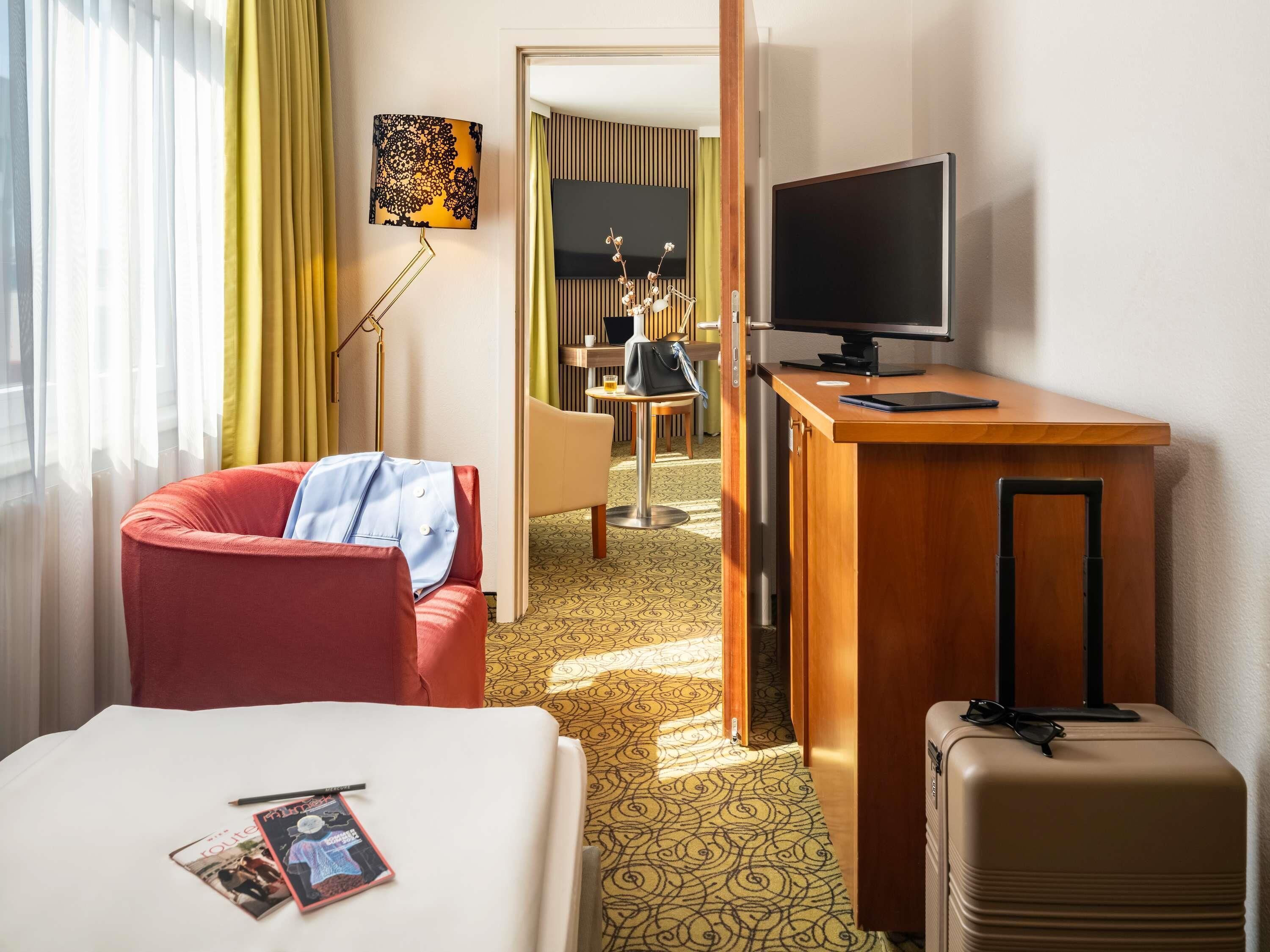 A hotel room with a bed, a desk with a TV, a red chair, a suitcase, and a lamp.