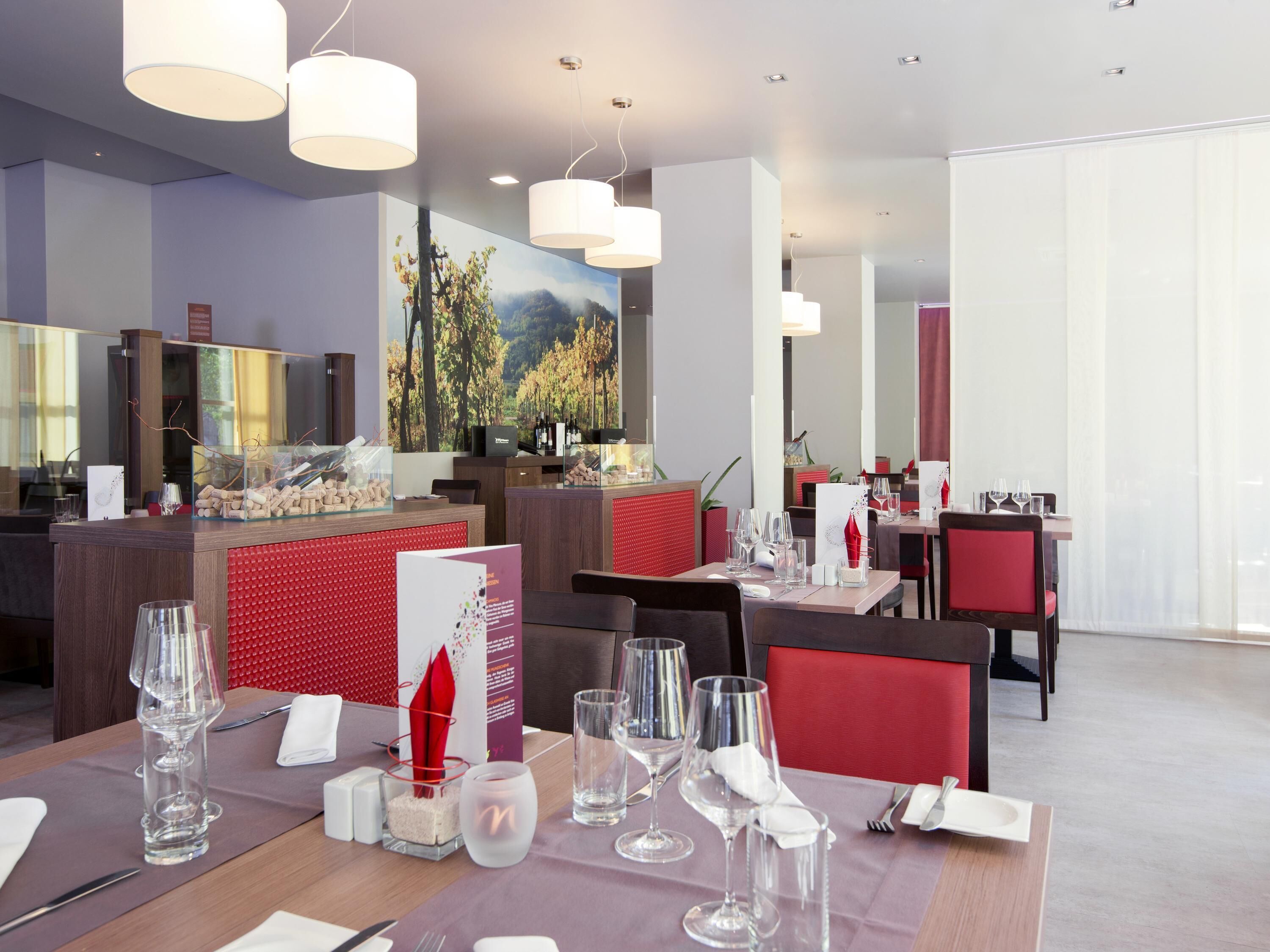 A modern restaurant with a red and white color scheme, featuring a dining area with tables set for service, a bar area with a wine display, and a large window with a view of trees.