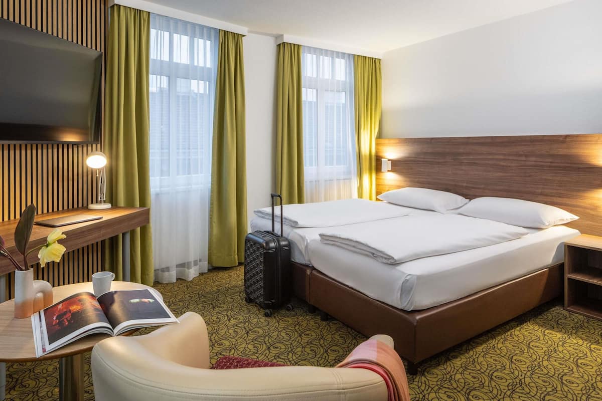 Ein Hotelzimmer mit Bett, Schreibtisch mit Lampe, Stuhl, Fernseher und Koffer.