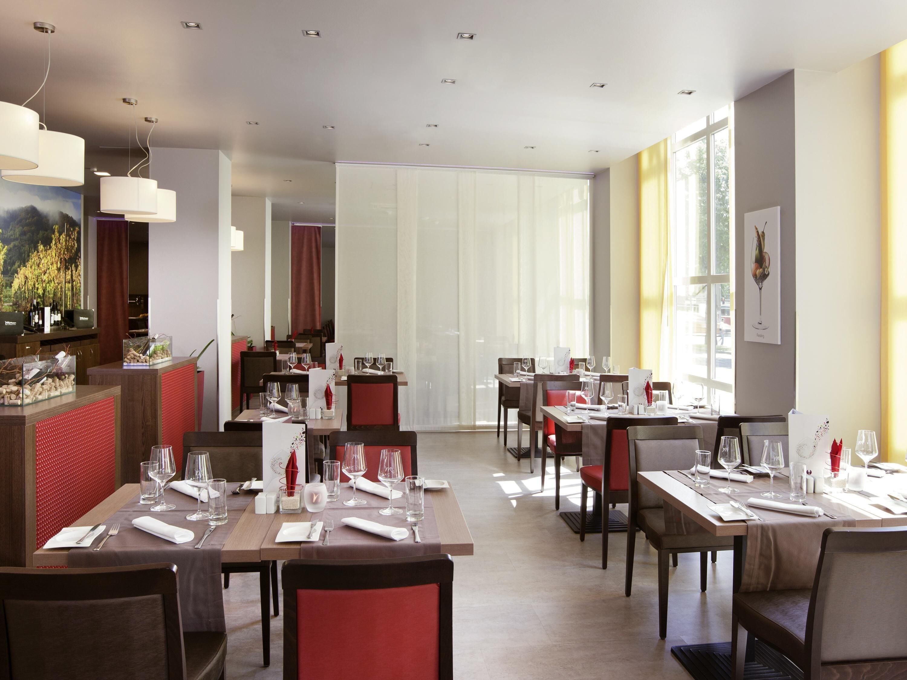 Un restaurant moderne doté de plusieurs tables, de chaises rembourrées de couleur rouge et d’un bar où sont exposées des bouteilles de vin.