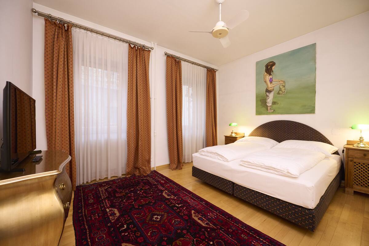 A bedroom with a bed, a TV, a desk, a chair, a rug, and a painting on the wall.