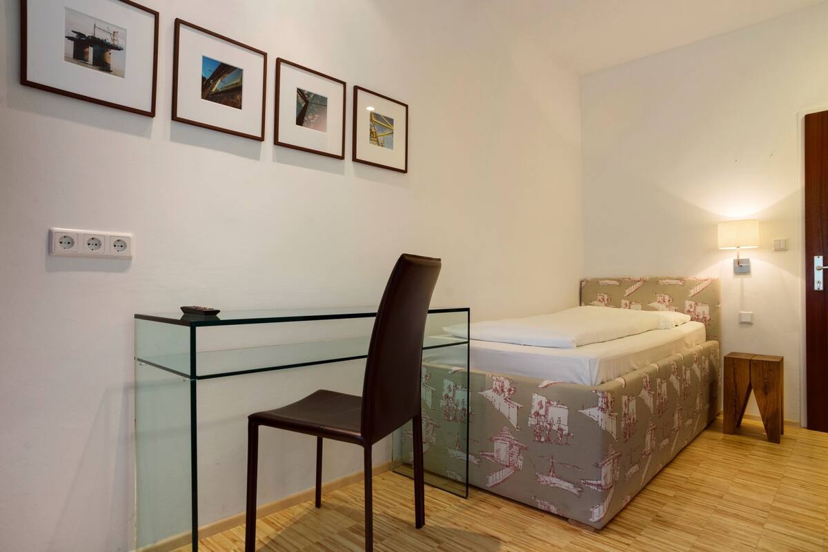 A bedroom with a bed, a desk with a chair, and a wall-mounted shelf.