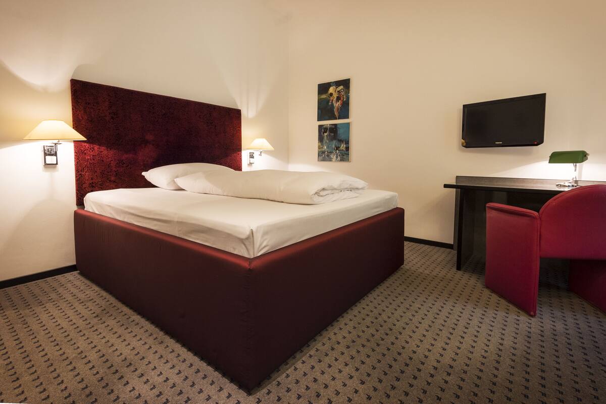 A hotel room with a large bed, two bedside lamps, a flat-screen TV, a desk, and a red chair.