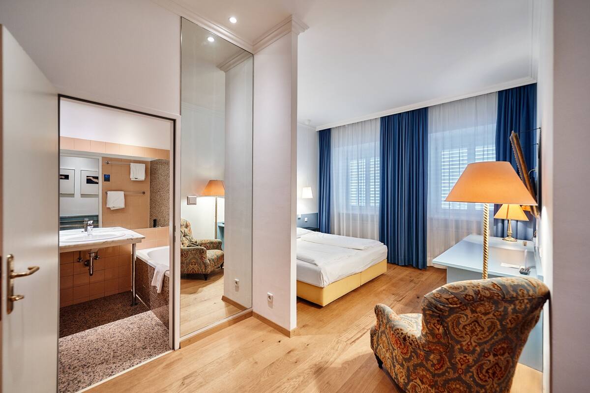A hotel room with a bathroom, a bed, a chair, and a desk with a lamp.