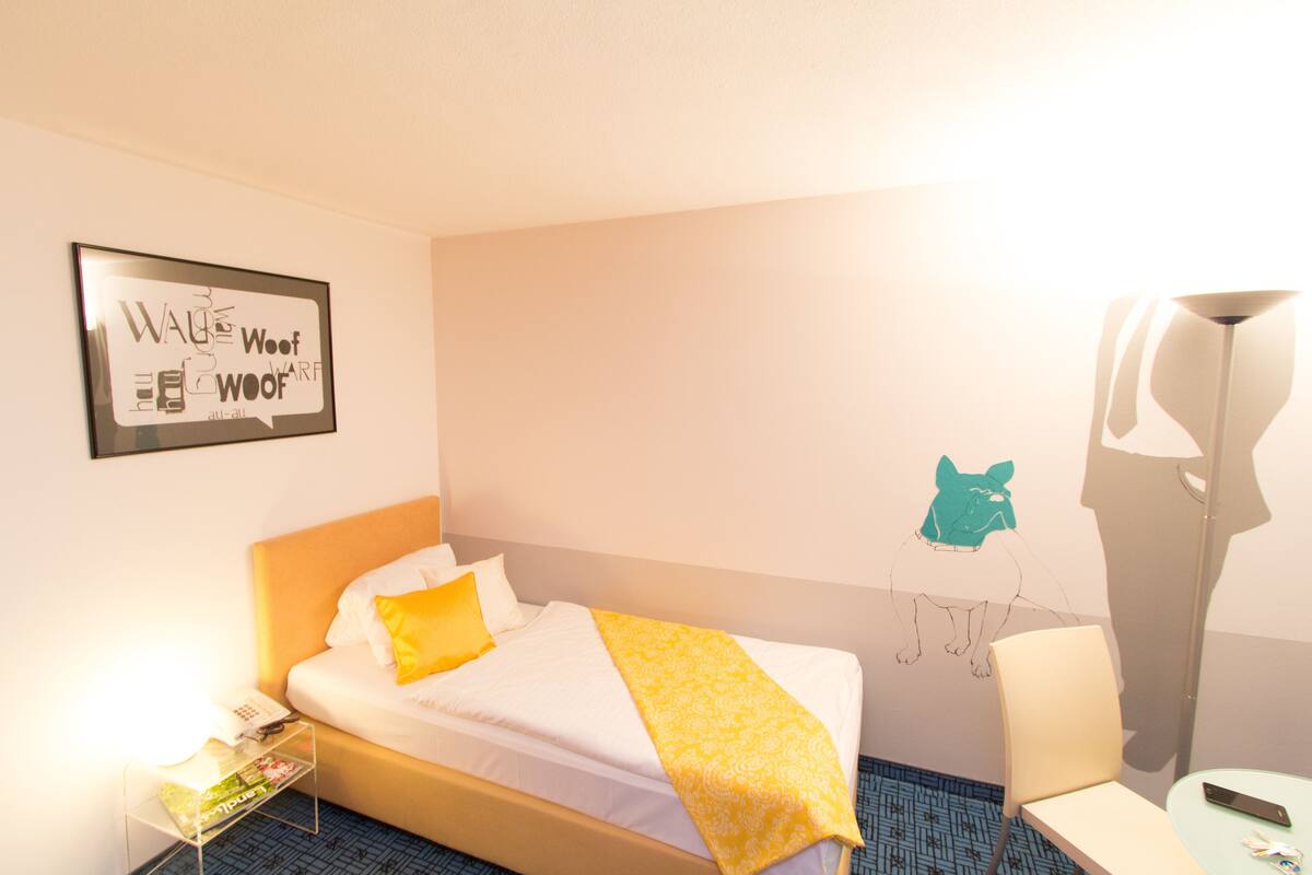 A hotel room with a bed, a bedside table with a phone and magazines, a chair, a small table with a remote control, and a wall art piece.