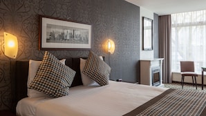 A hotel room with a large bed, two wall-mounted lamps, a framed cityscape picture, a fireplace, and a window with curtains.