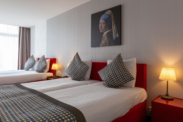 A hotel room with two beds, red headboards, patterned pillows, a painting on the wall, and a lamp on the nightstand.