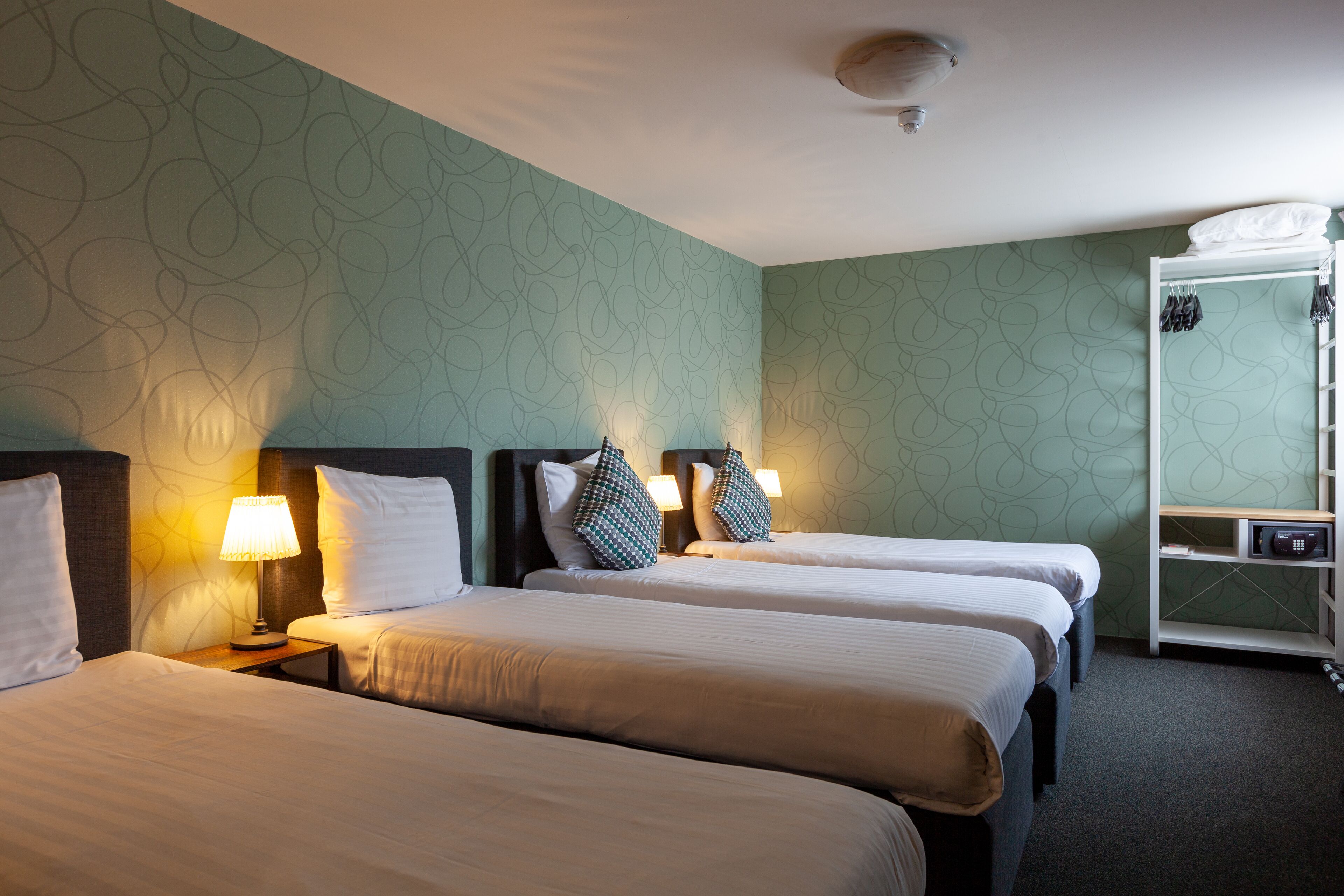 A hotel room with three beds, a wardrobe, and a small table with a lamp.