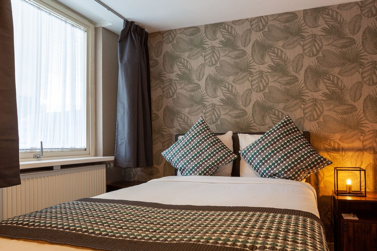 A neatly made bed with patterned pillows, a bedside table with a lamp, and a window with curtains.