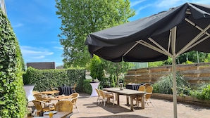 An outdoor patio with wicker furniture and a large umbrella, surrounded by greenery and a brick floor.