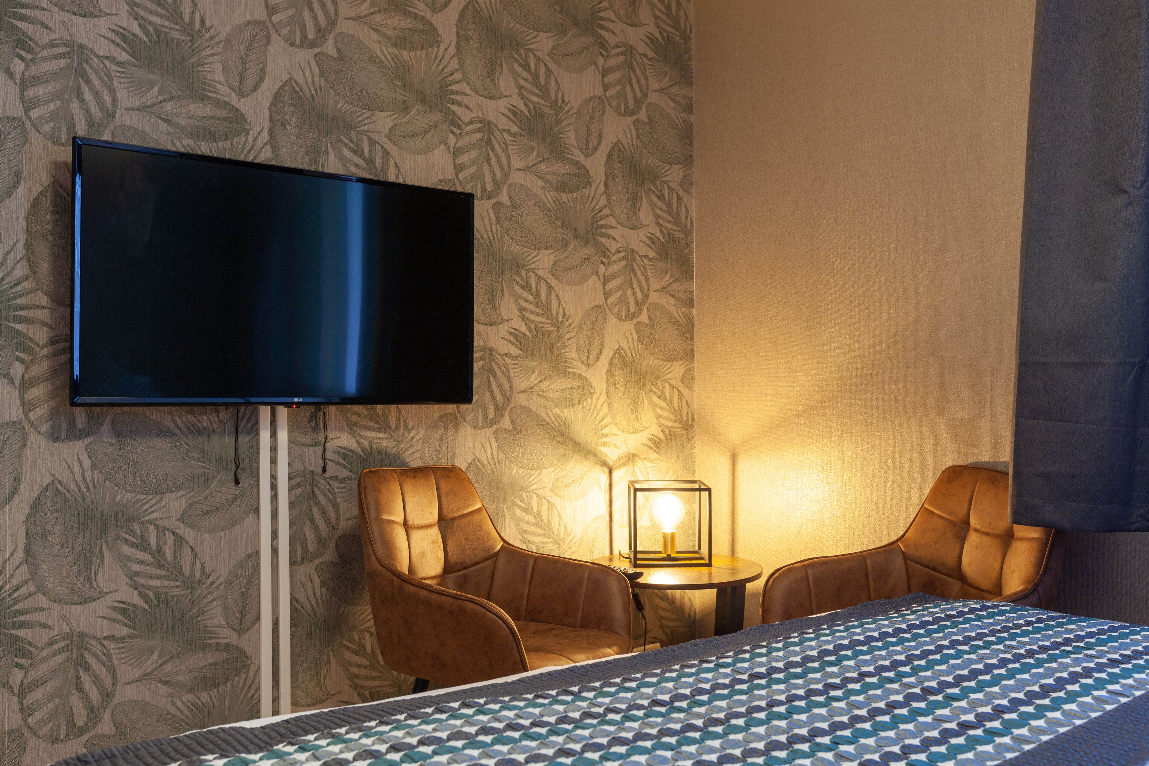 Ein Hotelzimmer mit einem Bett, einem an der Wand befestigten Fernseher, zwei Ledersesseln, einem kleinen Tisch mit einer Lampe und einer wanddekorierten Tapete.