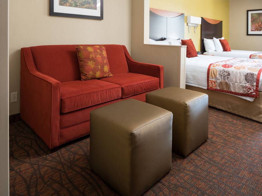 Ein Hotelzimmer mit einem roten Sofa, zwei Hocker, einem Bett mit gemustertem Überwurf und einem an der Wand montierten Fernseher.