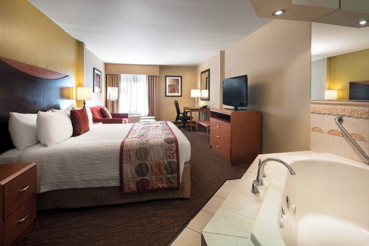 Habitación de hotel con una cama grande, un escritorio con silla, televisión, baño con bañera y espejo.