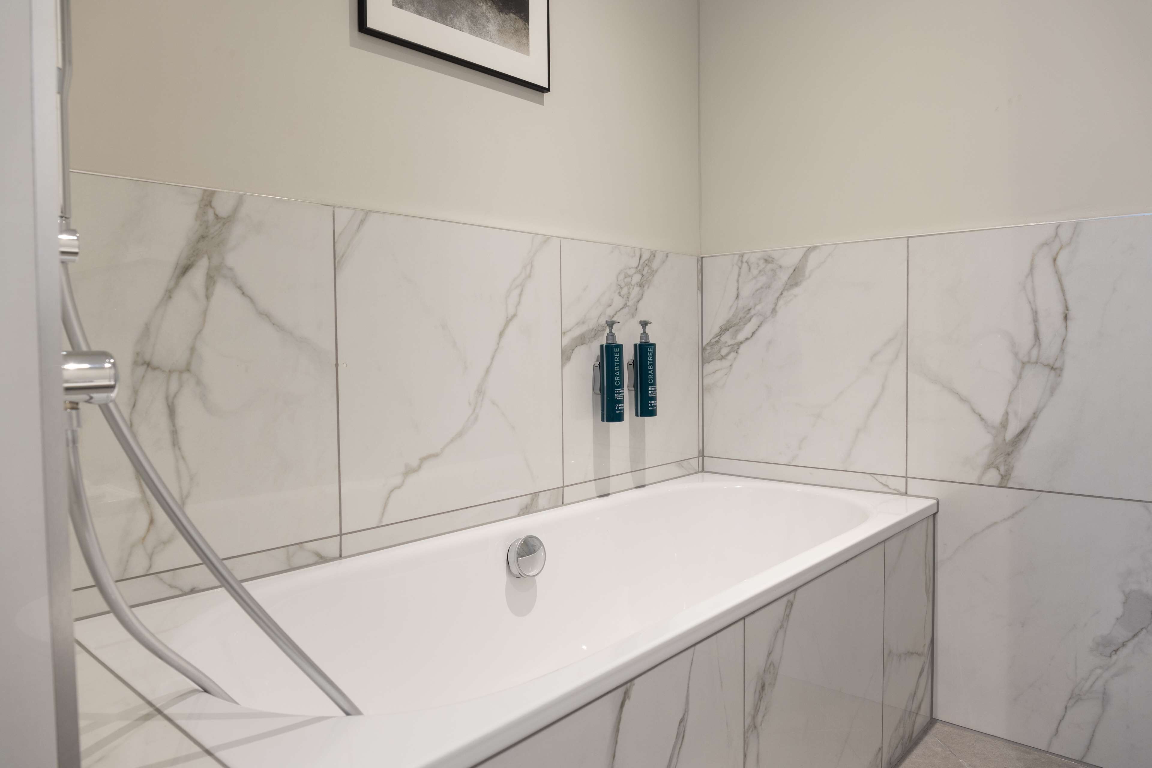 A modern bathroom with a marble-tiled wall, a white bathtub, and two blue toiletry bottles on the wall.