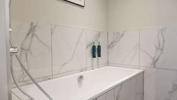 A modern bathroom with a marble-tiled wall, a white bathtub, and two blue toiletry bottles on the wall.