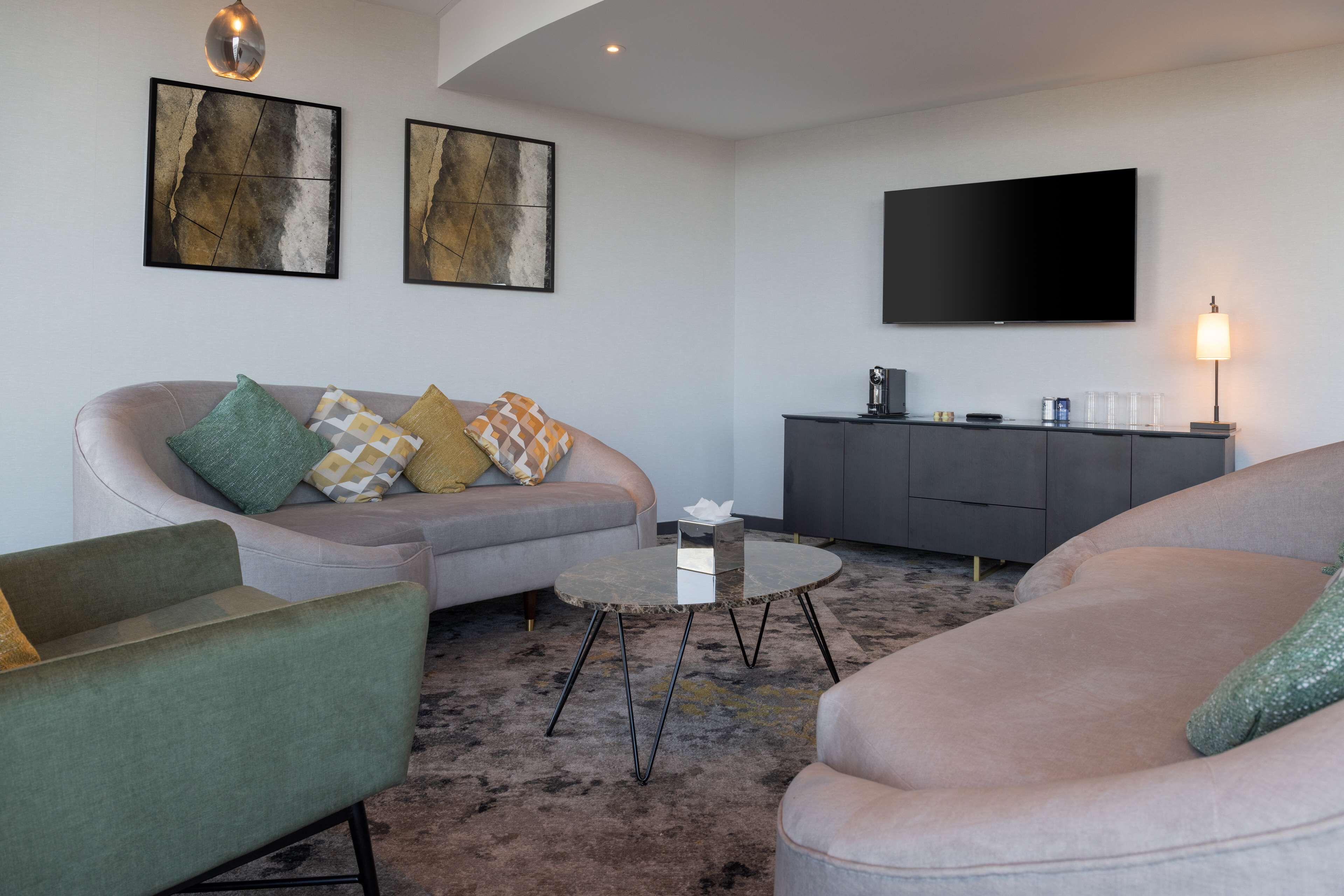 Ein modernes Wohnzimmer mit einem grauen Sofa, einem grünen Sessel und einem runden Couchtisch. An der Wand ist ein Flachbildfernseher angebracht und daneben ein Schrank mit einer Lampe.