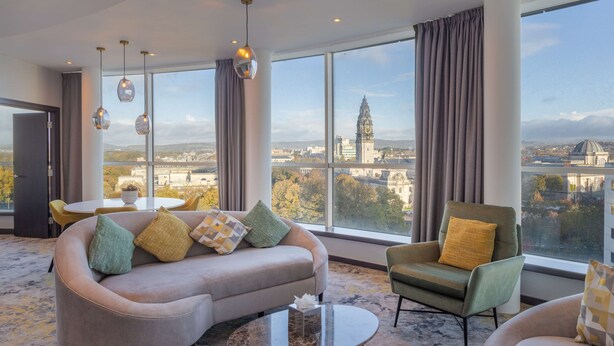 A modern hotel room with a large window offering a city view, a sofa with patterned cushions, a green armchair, and pendant lighting. - Hilton Cardiff (Cardiff)