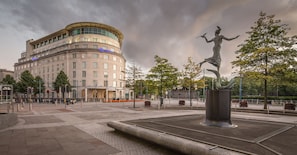 A hotel with a curved facade, a statue in the foreground, and a paved plaza with benches and trees.