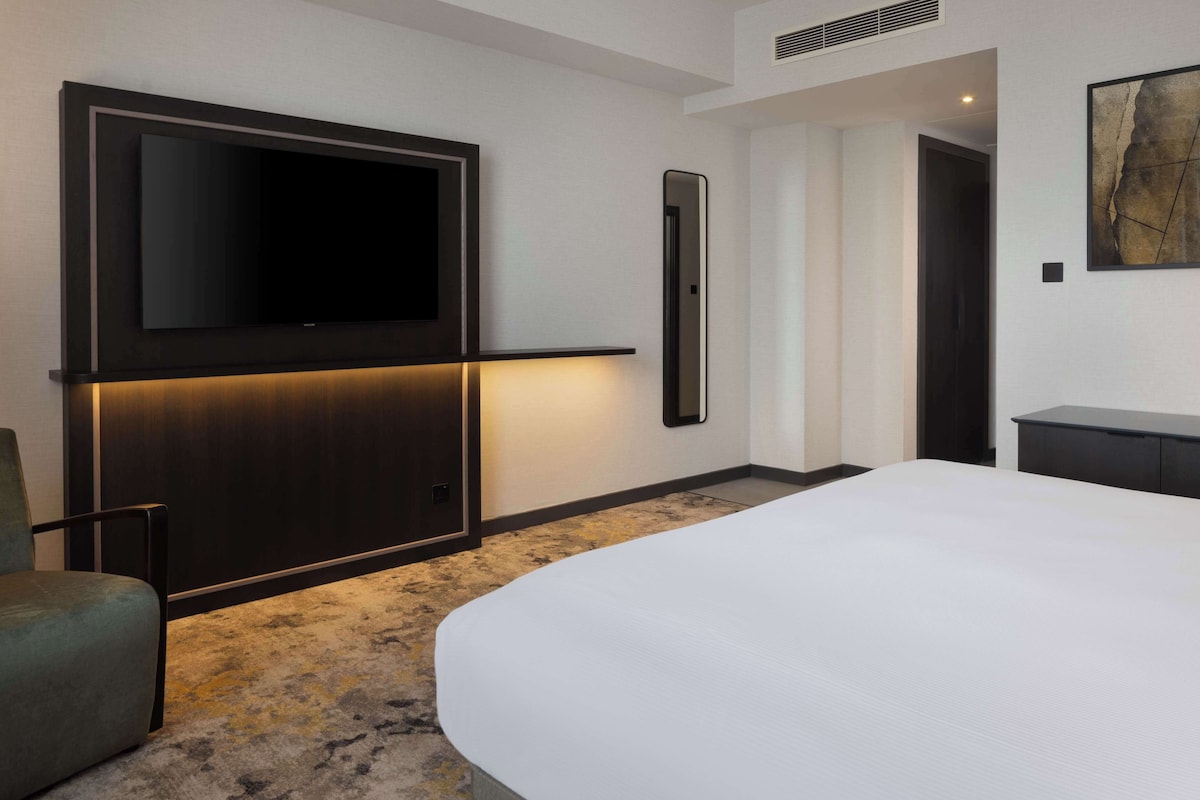 A modern hotel room with a large bed, a flat-screen TV mounted on the wall, a bedside table, and a carpeted floor.