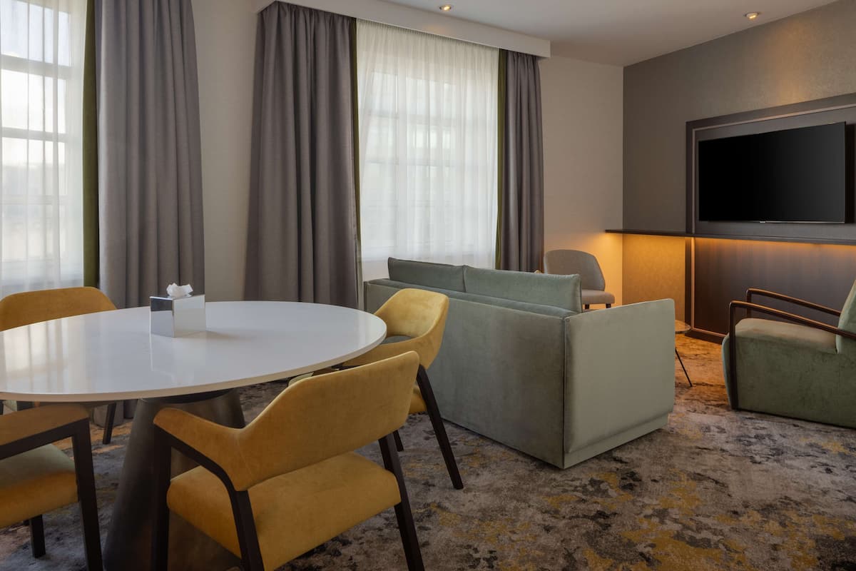A modern hotel room with a round table, yellow chairs, a grey sofa, and a flat-screen TV mounted on the wall.