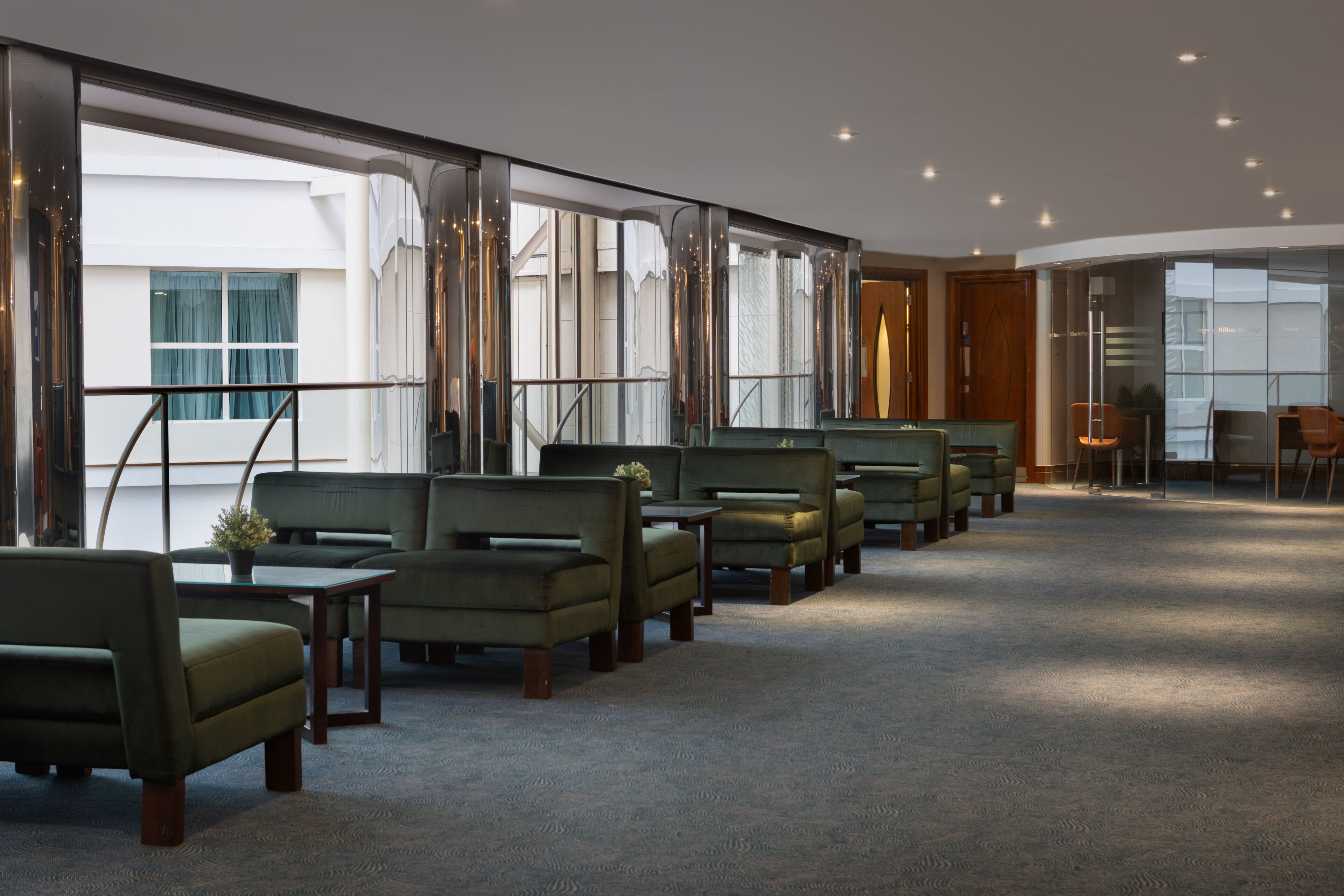 A hotel lobby with a row of green armchairs, a carpeted floor, and large windows offering a view of the building's exterior.