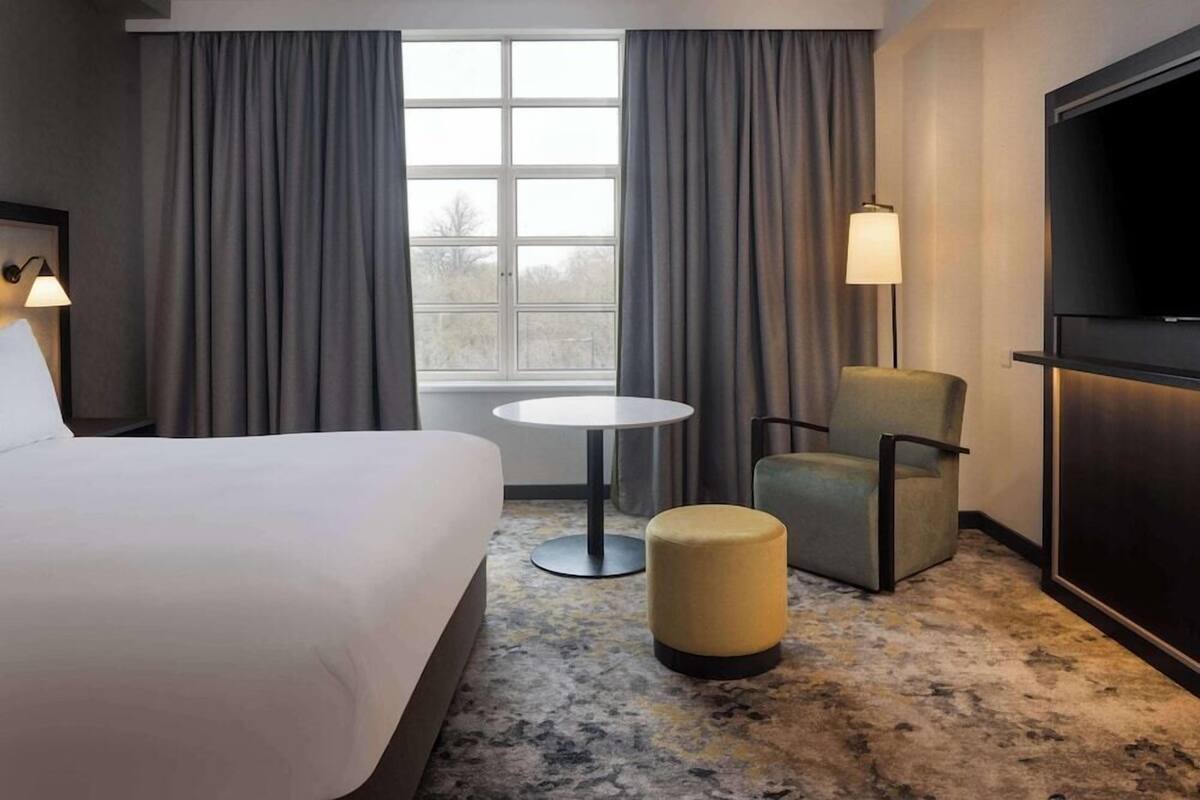 A hotel room with a bed, a round table, a chair, a small ottoman, a TV, and a window with curtains.