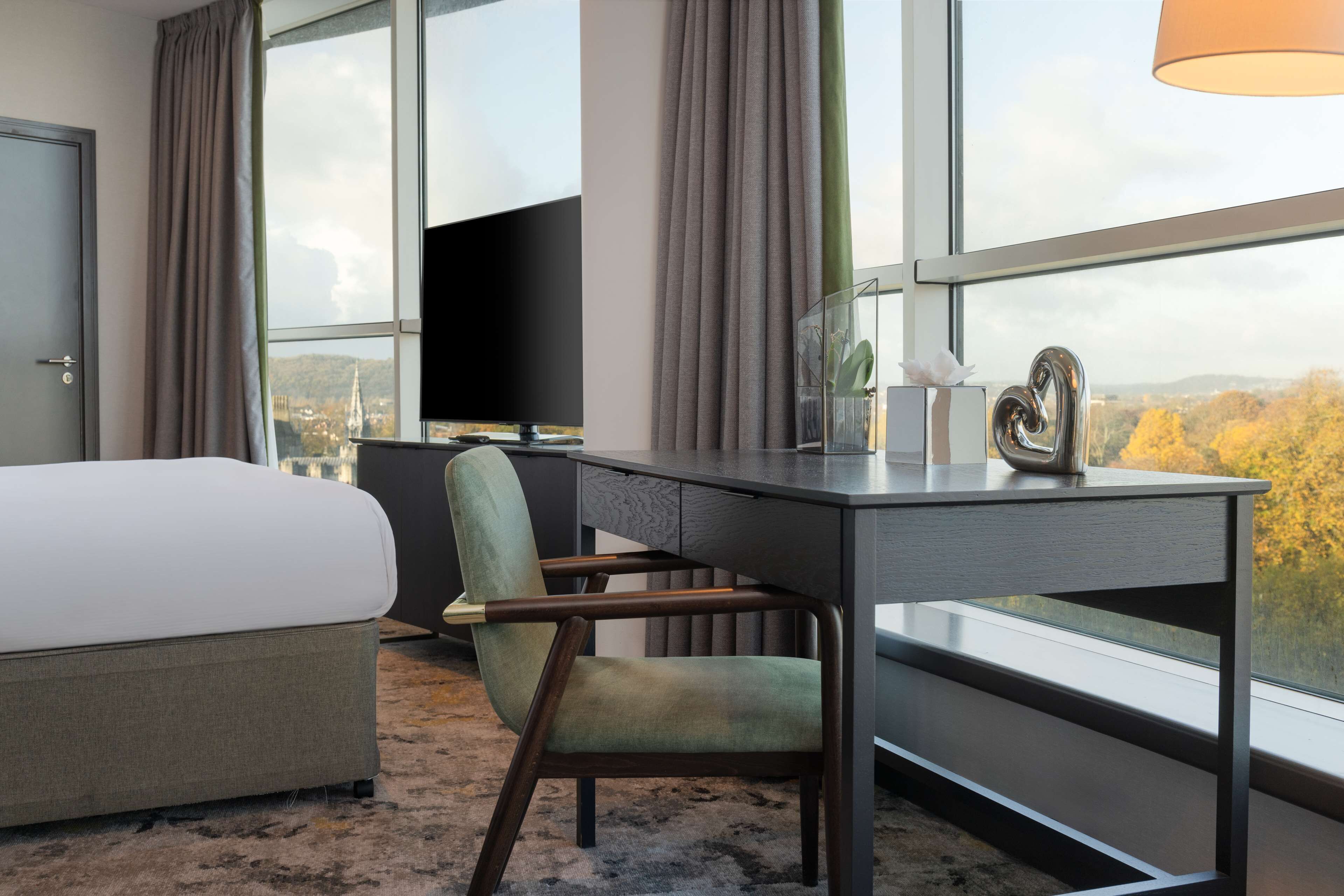 A hotel room with a bed, a desk, a chair, and a large window offering a view of a cityscape and autumn foliage.