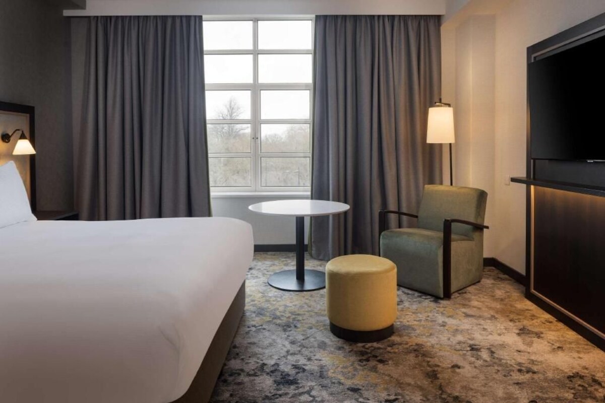 Una habitación de hotel con una cama, una mesa redonda, una silla, un reposapiés, un televisor y un ventanal con cortinas.