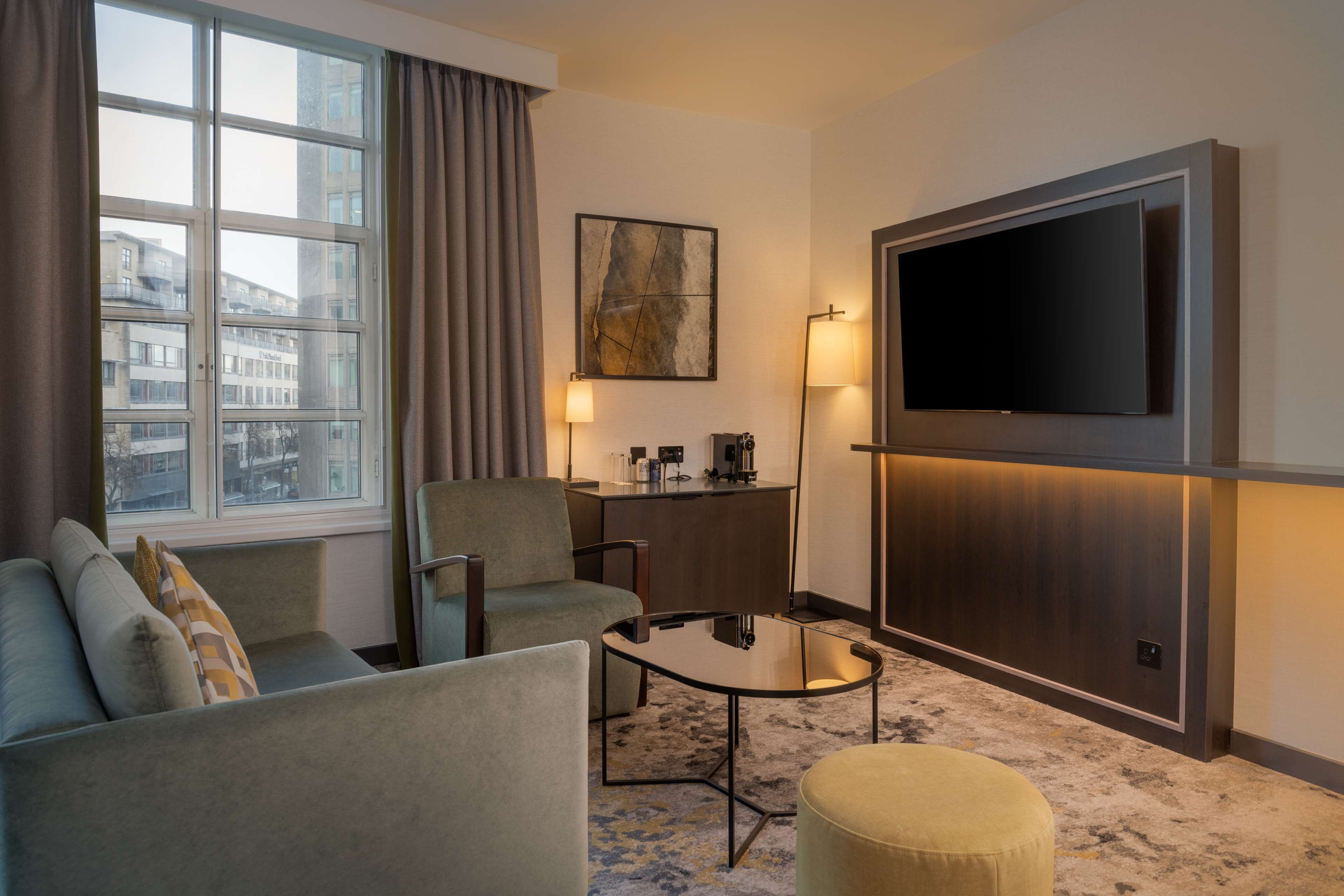 A hotel room with a sofa, armchair, small table, ottoman, TV, desk, lamp, and a window with curtains.