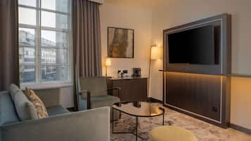 A hotel room with a sofa, armchair, small table, ottoman, TV, desk, lamp, and a window with curtains.