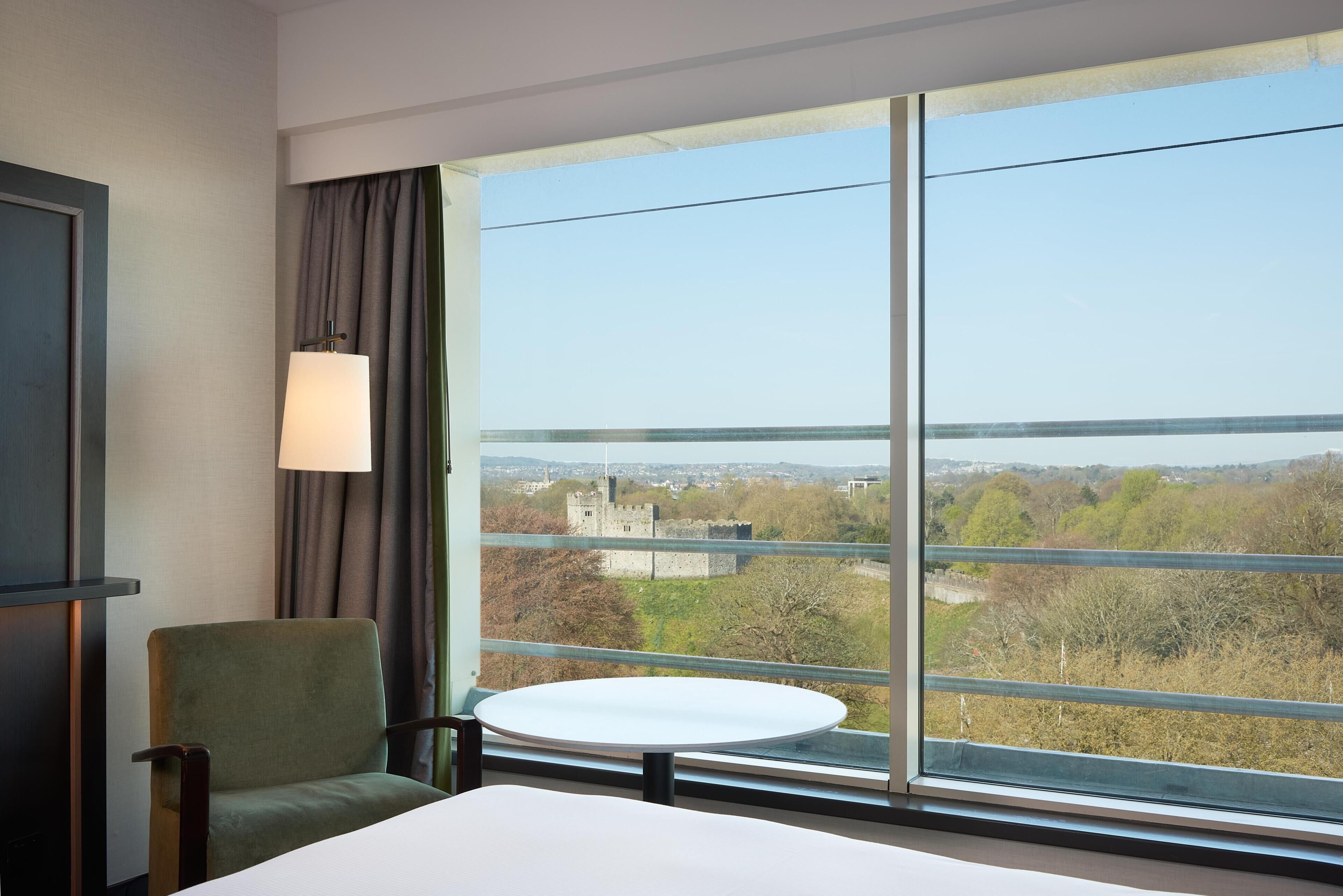 A hotel room with a large window, a round table, a green chair, and a lamp.