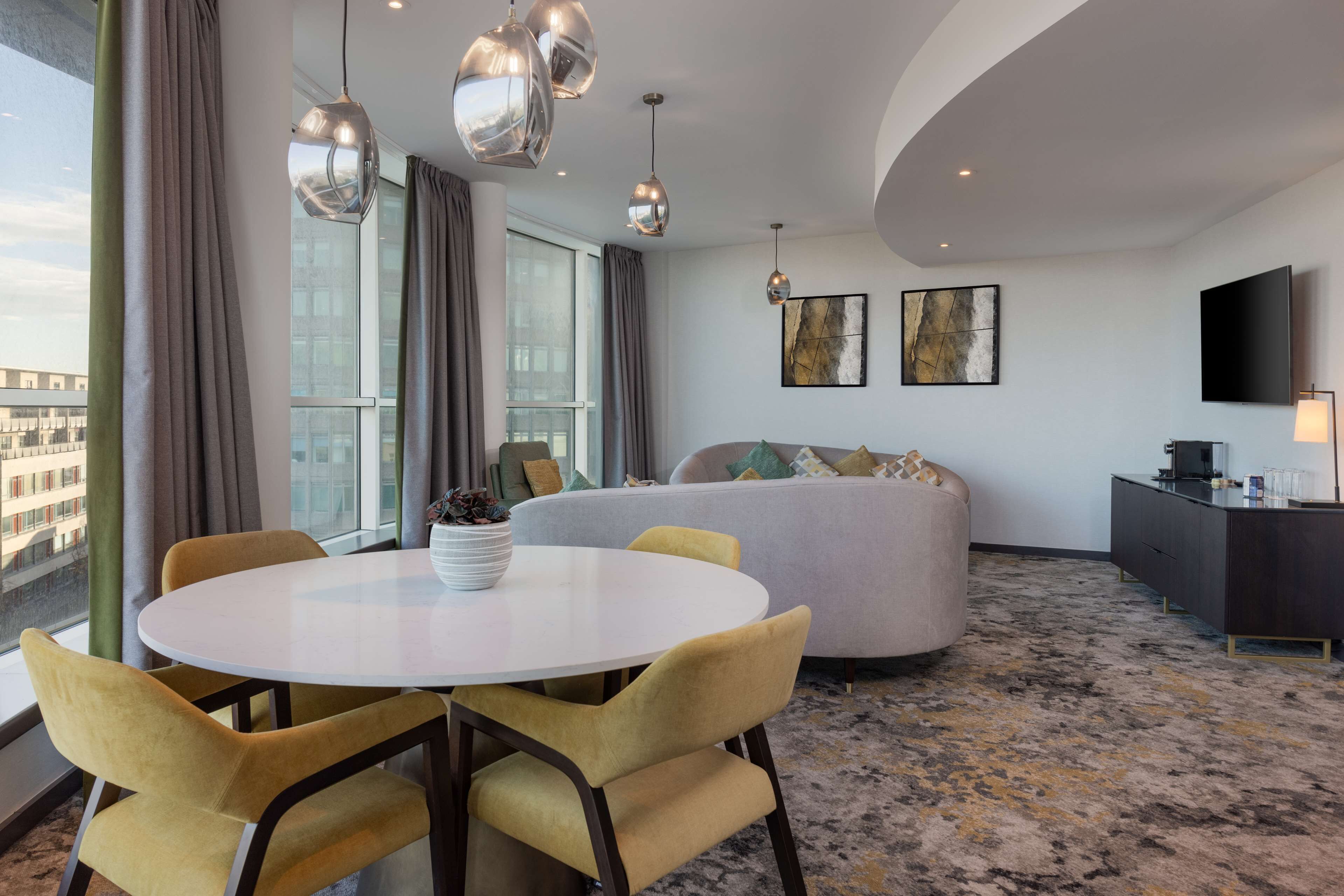 Een moderne hotelkamer met een ronde eettafel, gele stoelen, een grijze bank en een groot raam met uitzicht over de stad.
