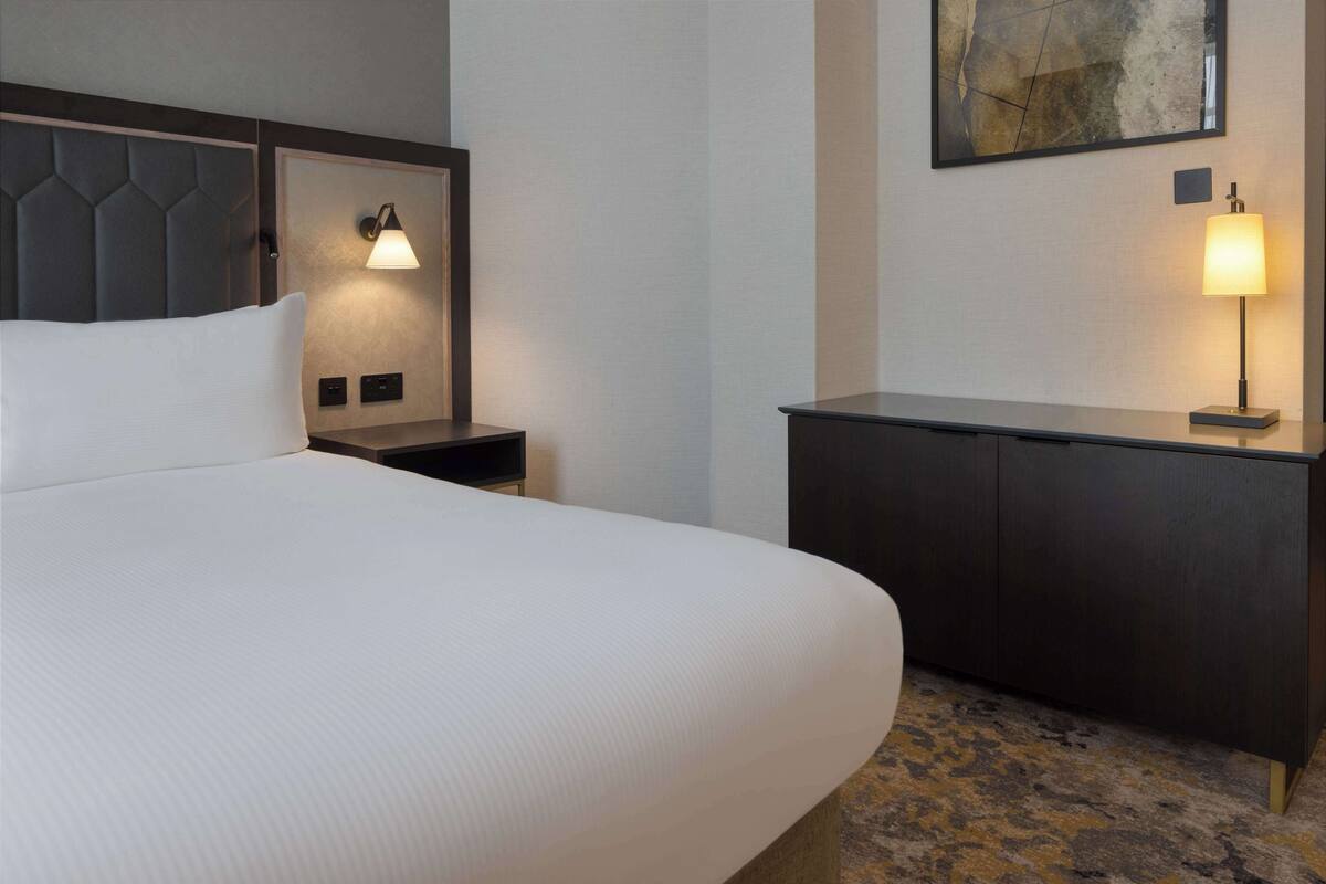 A hotel room with a large bed, a bedside table with a lamp, a small desk, and a painting on the wall.