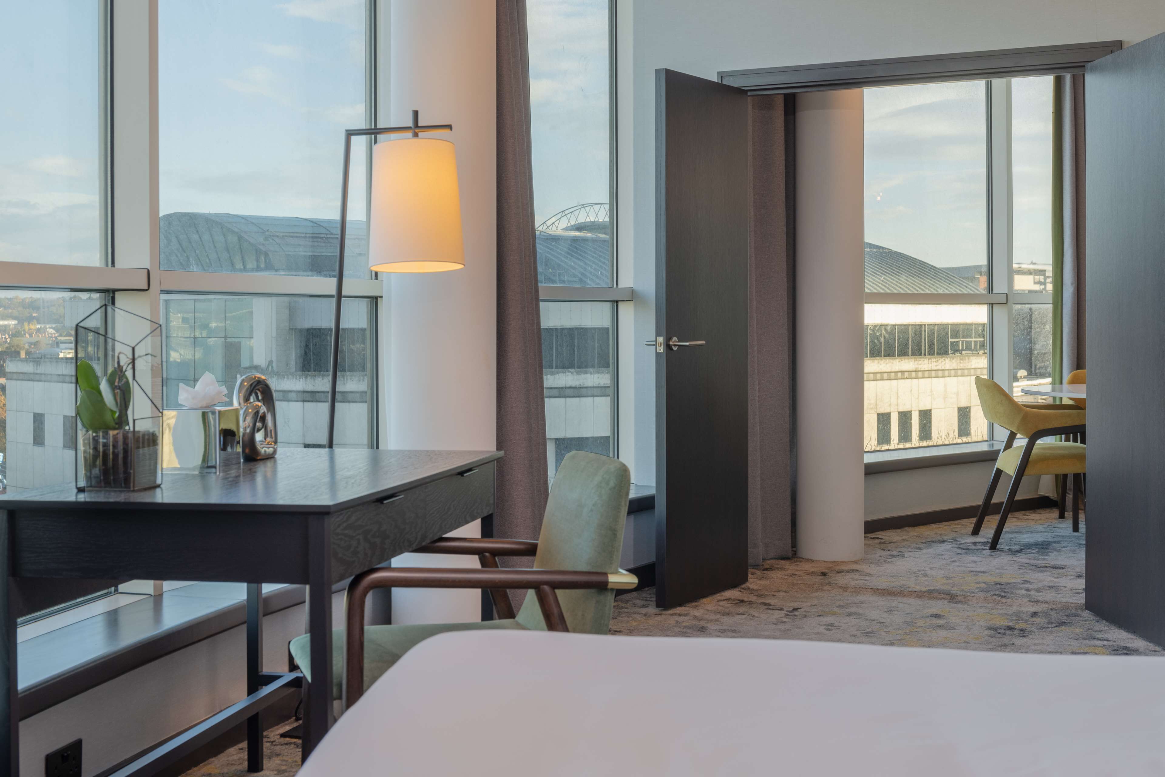 A hotel room with a large window, a desk with a lamp, a chair, and a bed.