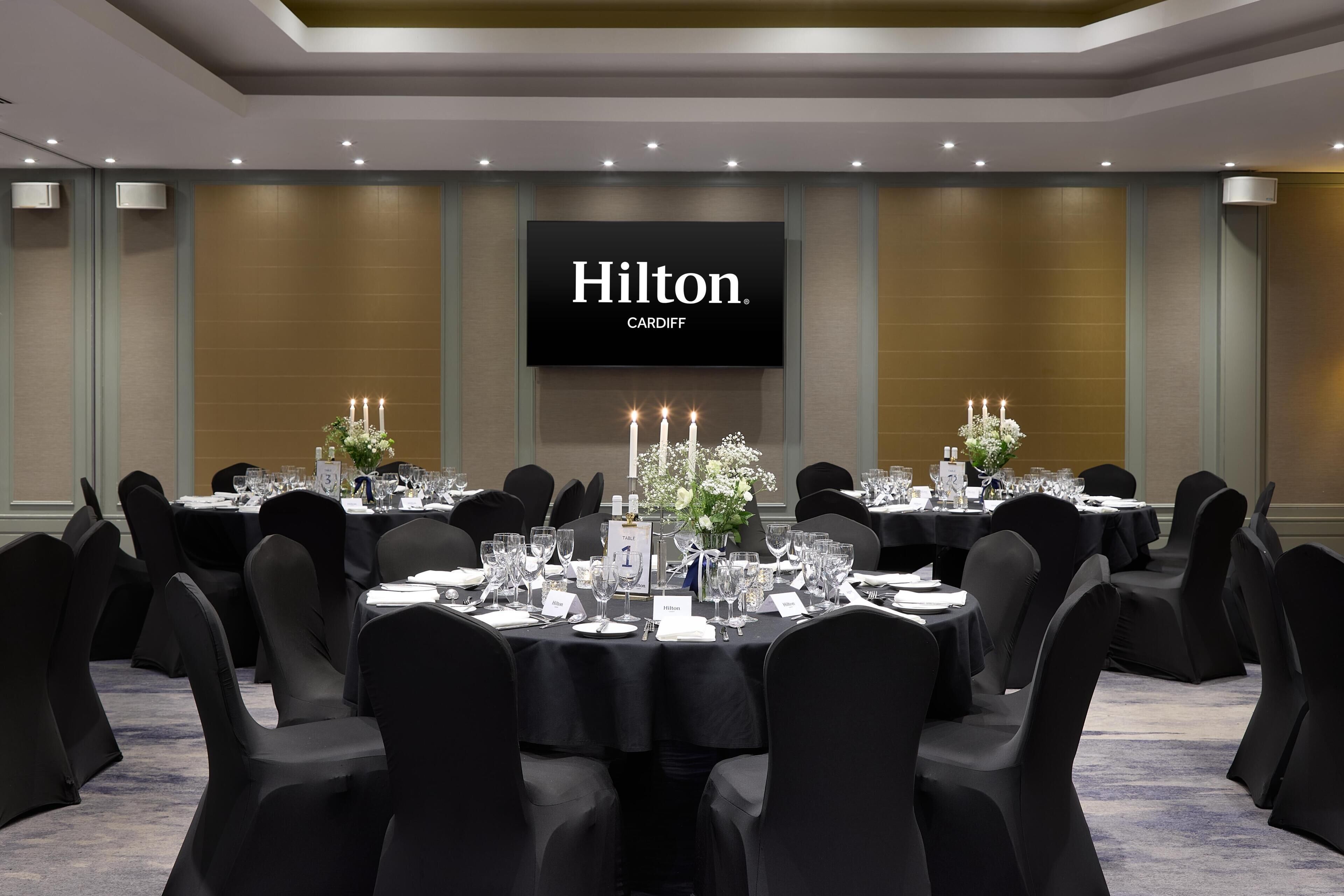 A conference room with round tables, black chairs, and a large screen displaying 'Hilton Cardiff'.