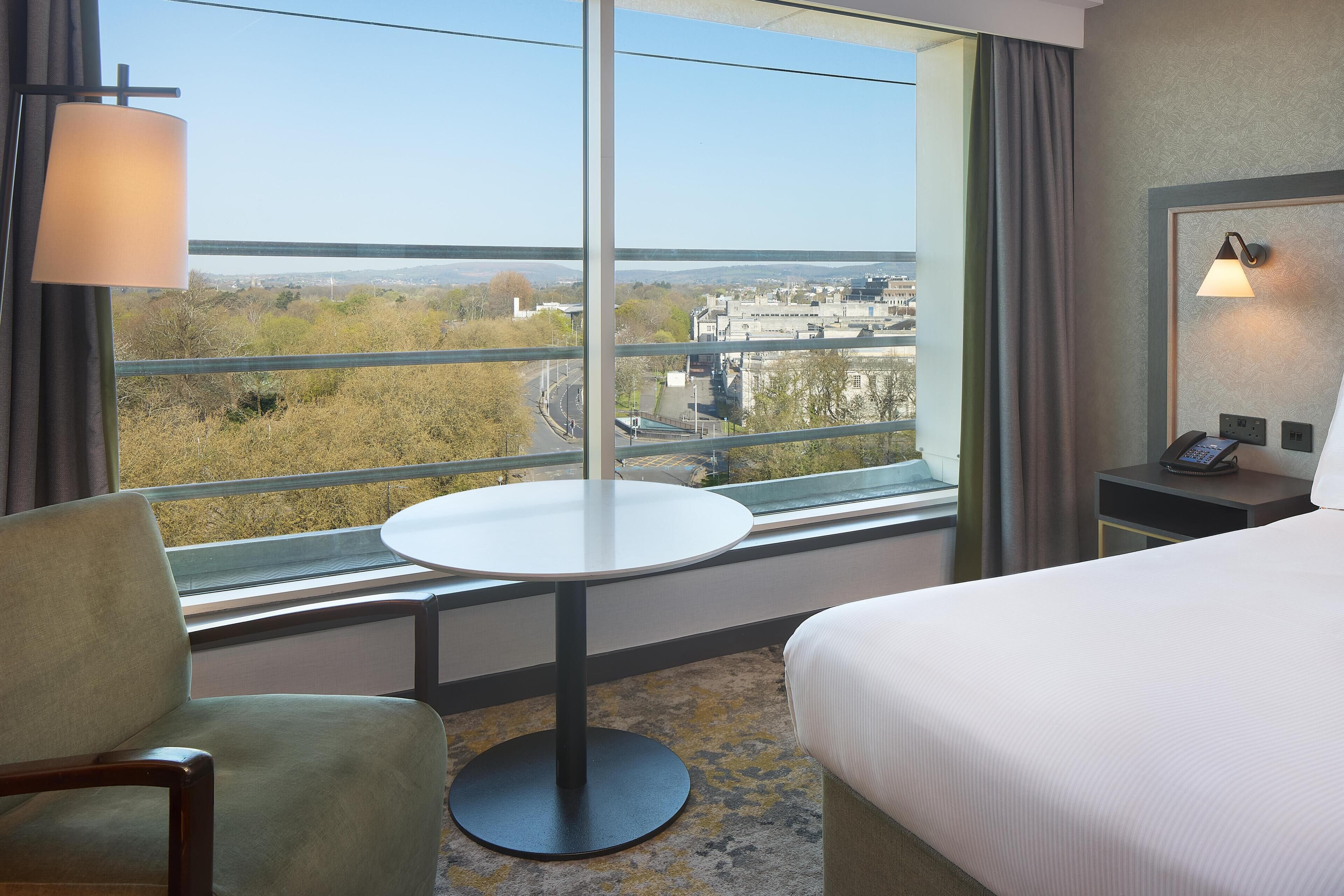A hotel room with a bed, a round table, a chair, a nightstand with a phone, and a large window offering a city view.