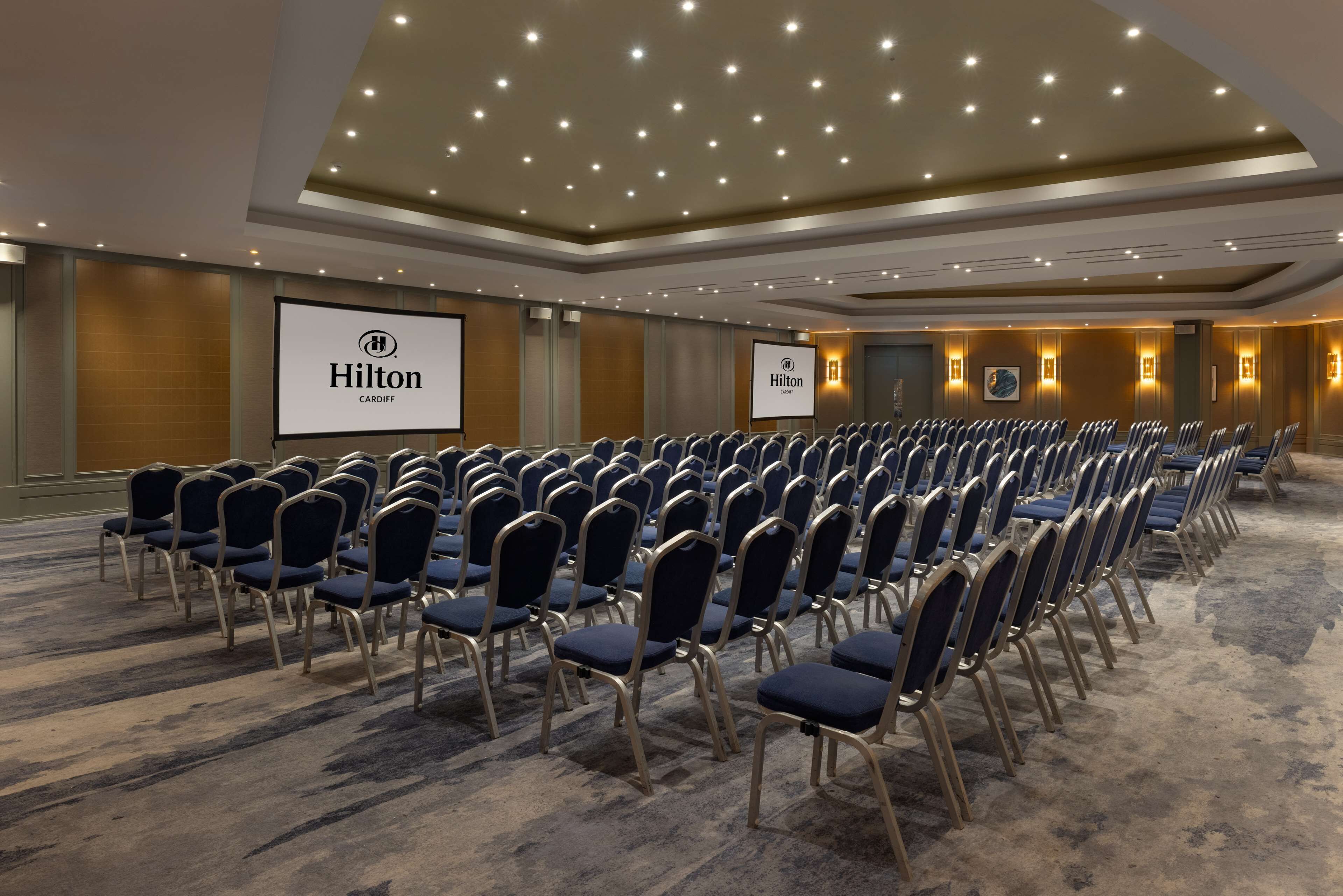 A conference room with rows of blue chairs, a large screen, and a "Hilton Cardiff" sign.