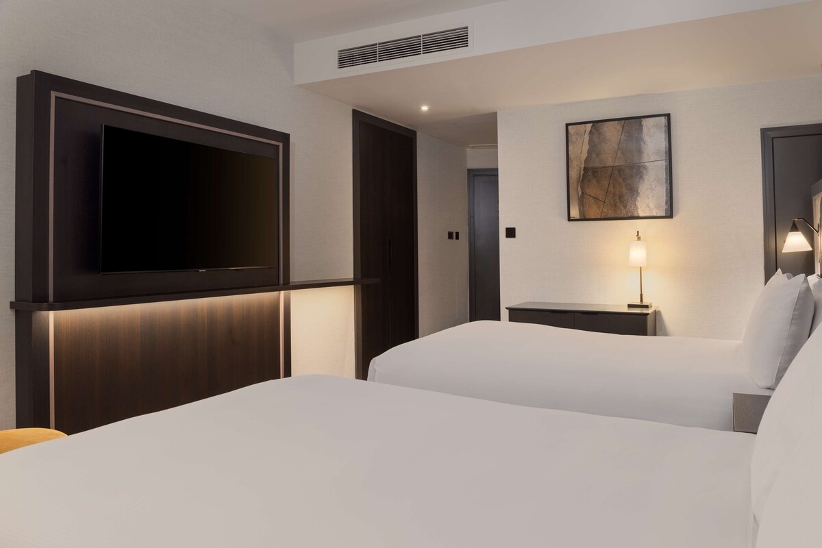 Habitación de hotel con un televisor de pantalla plana grande montado en la pared, dos camas con sábanas blancas, una mesita de noche con lámpara y un cuadro en la pared.