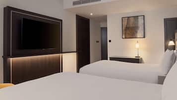 Habitación de hotel con un televisor de pantalla plana grande montado en la pared, dos camas con sábanas blancas, una mesita de noche con lámpara y un cuadro en la pared.