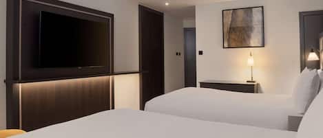 A hotel room with a large flat-screen TV mounted on the wall, two beds with white linens, a bedside table with a lamp, and a painting on the wall.