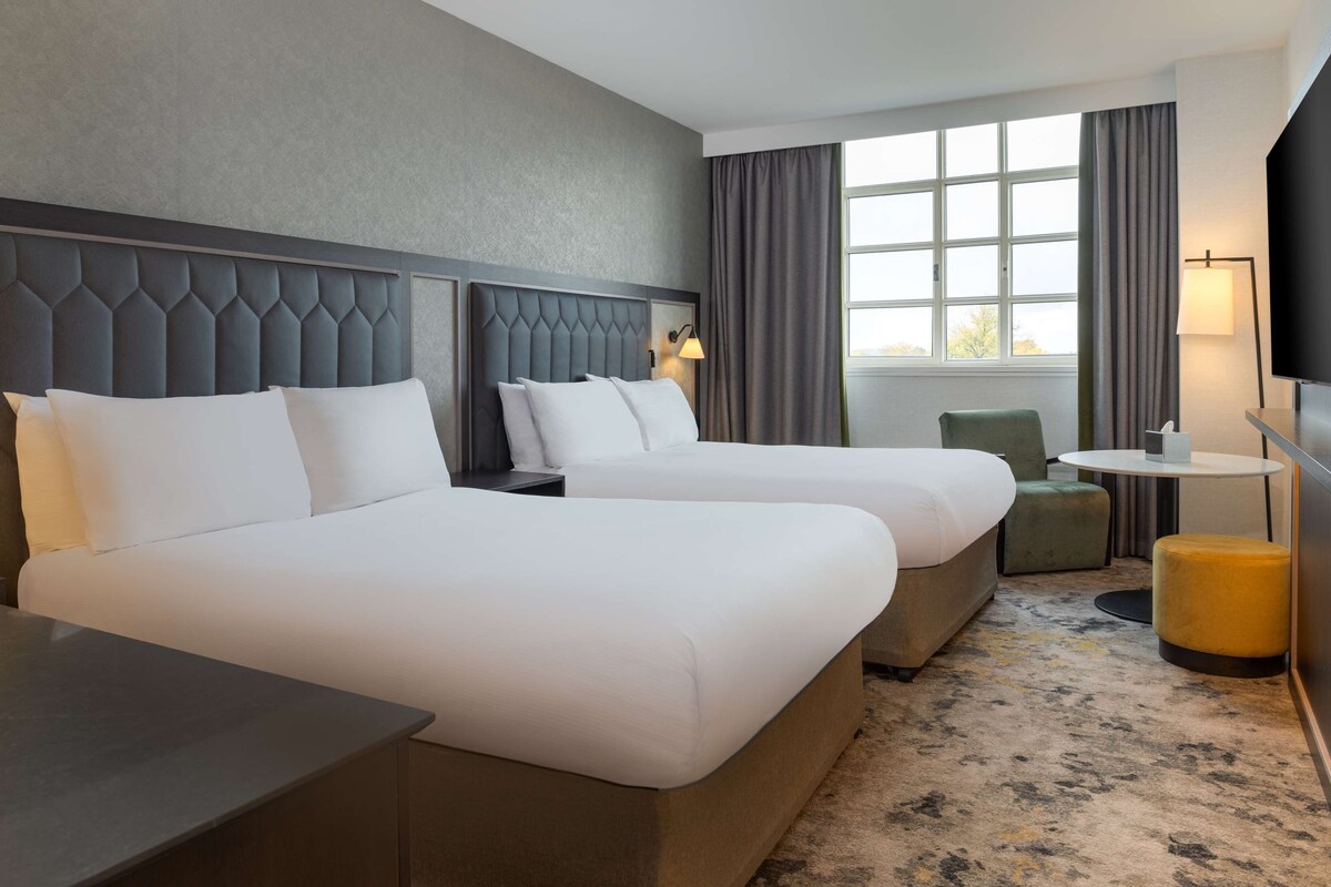 A hotel room with two beds, a small table, a chair, a TV, and a window with curtains.